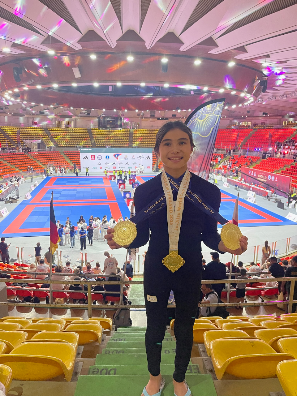 Bailey Heinsohn holding three gold medals above the competition floor at the 2025 JJIF World Championships in Bangkok while representing the Philippines.