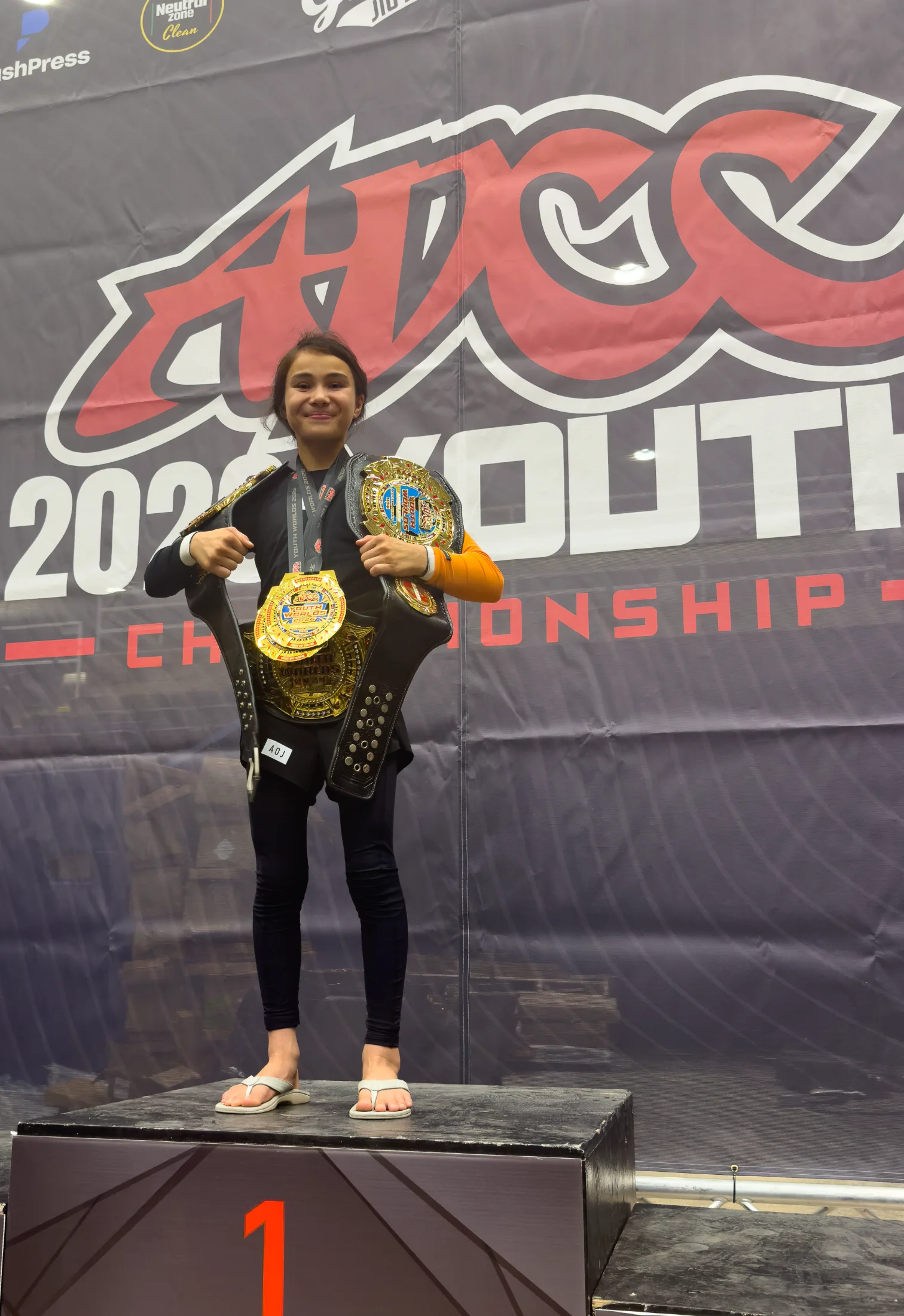 Bailey Heinsohn standing on the first-place podium with her championship belts and medal at the 2026 ADCC Youth World Championships in Austin, Texas.