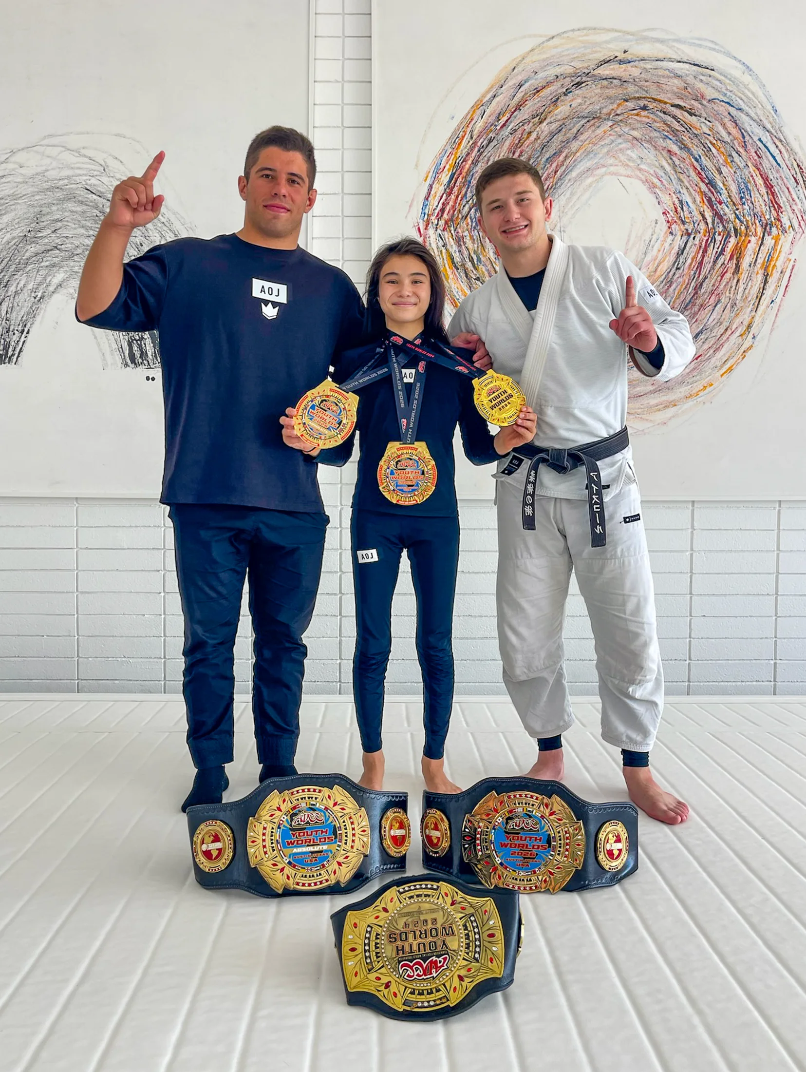 Bailey Heinsohn at Art of Jiu Jitsu Academy with Tainan Dalpra and Cole Abate, displaying her championship belts and medals after winning double gold at the 2026 ADCC Youth World Championships.
