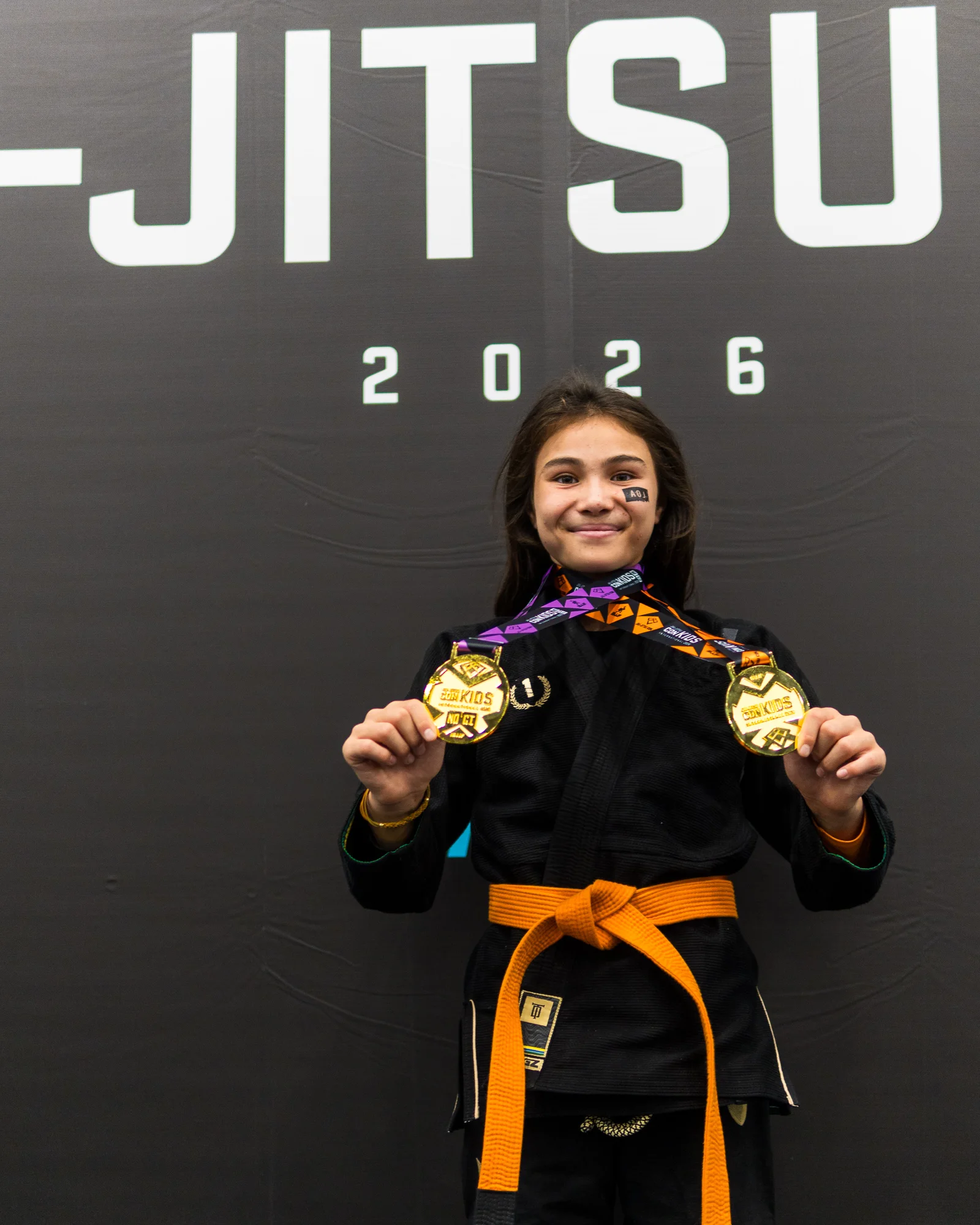 Bailey Heinsohn wearing an orange belt and black Gi while holding her Gi and No-Gi gold medals at the 2026 IBJJF Jiu-Jitsu CON in Las Vegas.