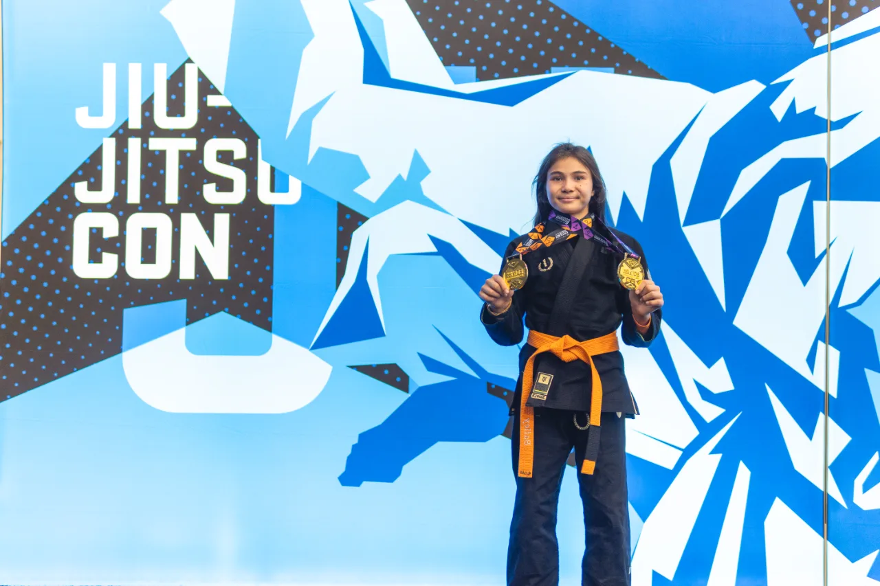 Bailey Heinsohn wearing her orange belt and holding two gold medals after winning Gi and No-Gi gold at the 2026 IBJJF Jiu-Jitsu CON Kids International in Las Vegas.